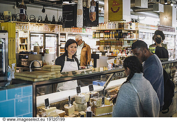 Smiling saleswoman looking at customers buying cheese from display cabinet at store