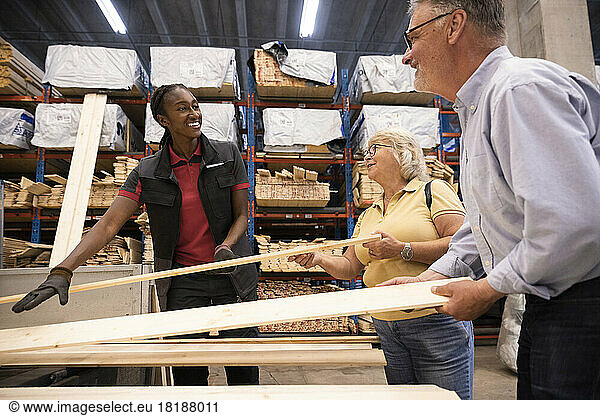 Smiling saleswoman looking at male and female customer holding planks at hardware store