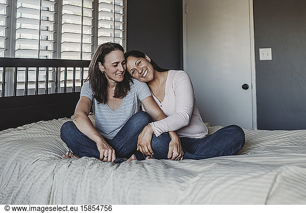 Smiling same sex couple snuggling on bed near window with shutters