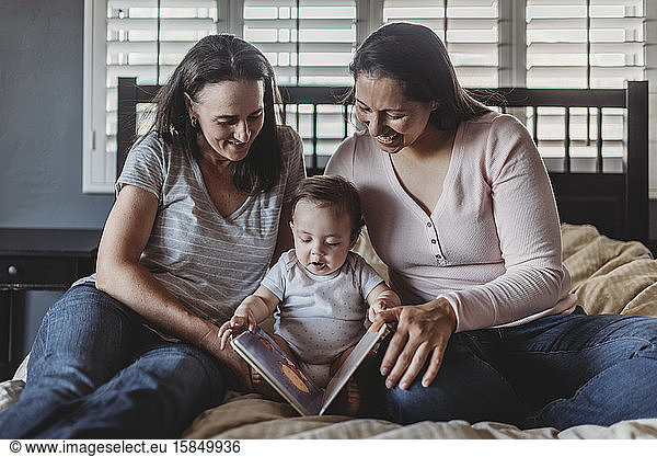 Smiling same sex parents reading with baby while sitting on bed