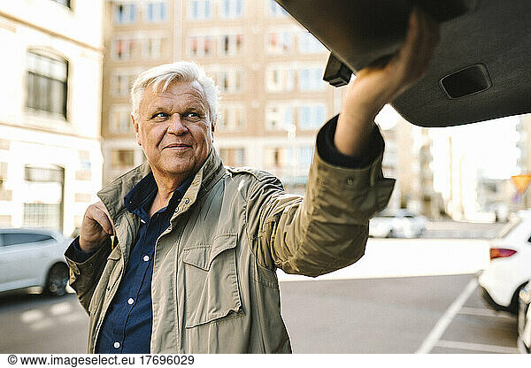 Smiling senior businessman closing car trunk while standing in parking lot