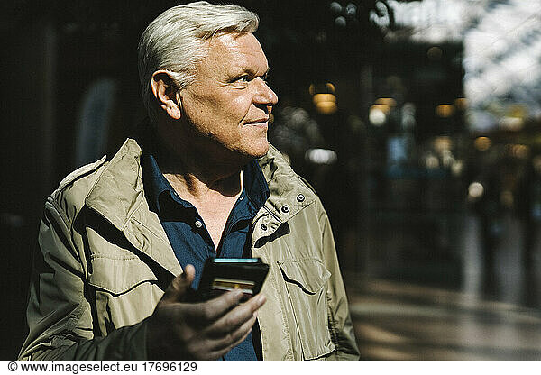 Smiling senior businessman contemplating while holding smart phone at railroad station