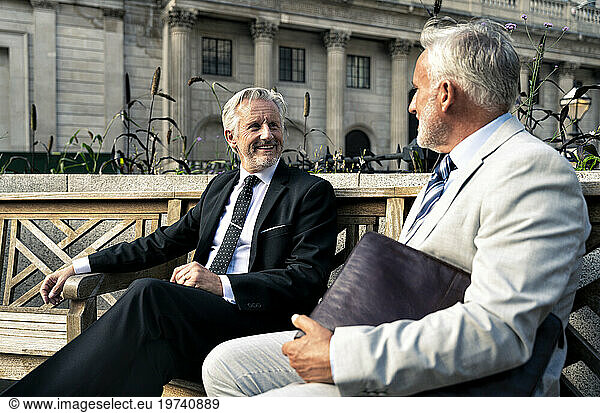Smiling senior businessmen having discussion together on bench in front of financial building