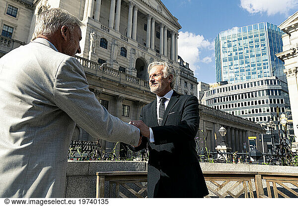 Smiling senior businessmen shaking hands together in front of financial buildings on sunny day