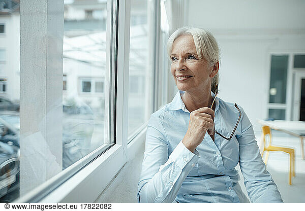 Smiling senior businesswoman holding eyeglasses looking through window in office