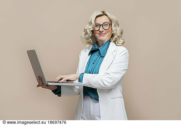 Smiling senior businesswoman holding laptop against colored background