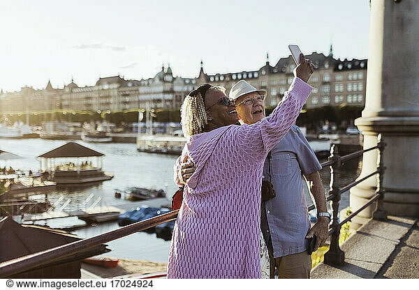 Smiling senior couple taking selfie while standing on bridge against lake in city