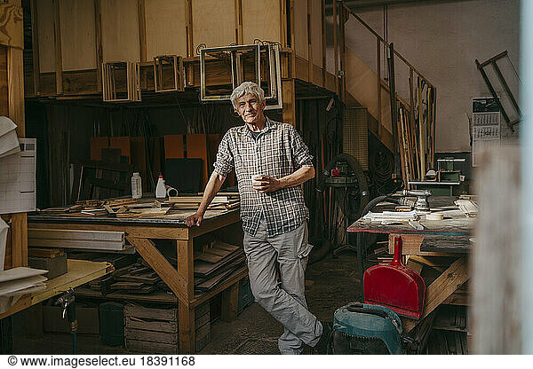Smiling senior craftsman leaning on workbench at carpentry workshop