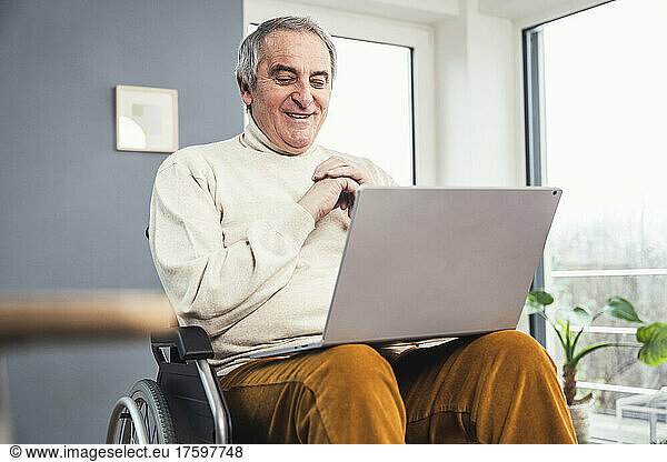 Smiling senior disabled man sitting with laptop on wheelchair at home