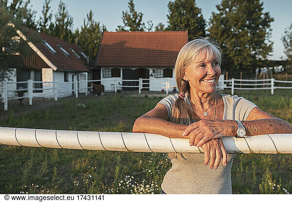 Smiling senior female farm worker leaning on fence at farm