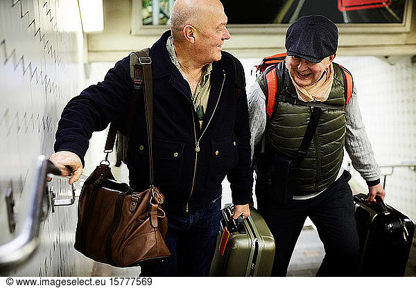 Smiling senior gay couple with luggage walking on staircase in subway