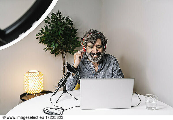 Smiling senior male podcaster with headphones using laptop at home office