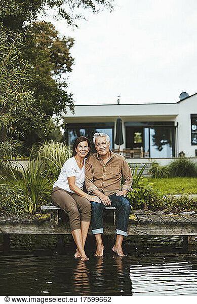 Smiling senior man and woman sitting on jetty by lake at garden