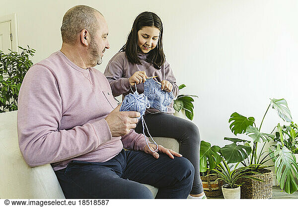 Smiling senior man assisting granddaughter in knitting sweater at home
