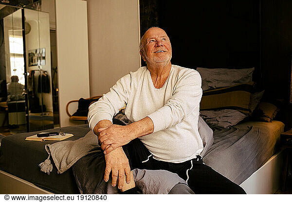 Smiling senior man looking up while sitting on bed at home