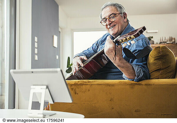 Smiling senior man playing guitar learning through tablet PC in living room