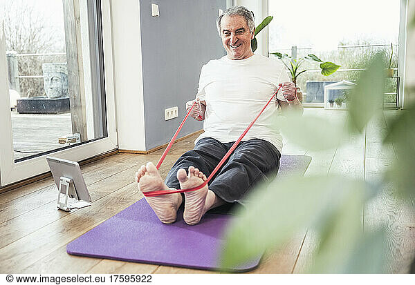 Smiling senior man pulling resistance band sitting on exercise mat at home