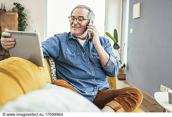 Smiling senior man talking on mobile phone looking at tablet PC in living room