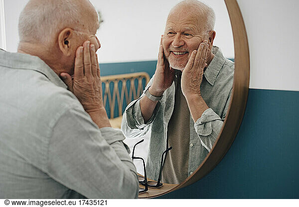 Smiling senior man touching skin while looking in mirror