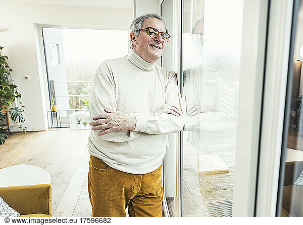 Smiling senior man with arms crossed looking through window at home