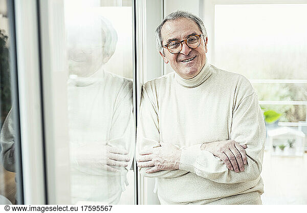 Smiling senior man with arms crossed standing by window at home