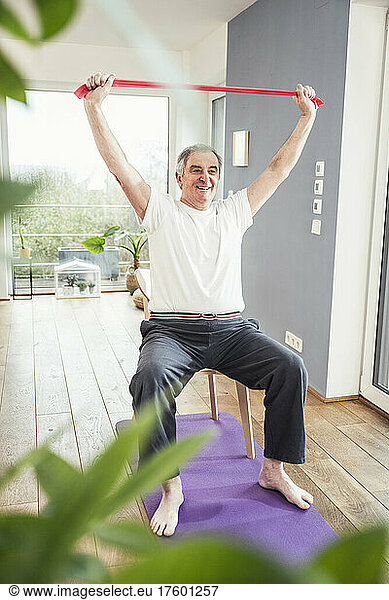 Smiling senior man with arms raised holding resistance band sitting on chair at home