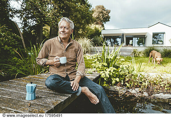 Smiling senior man with coffee cup sitting on jetty by lake at backyard