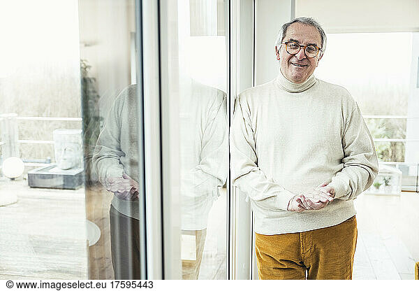 Smiling senior man with eyeglasses leaning on window at home