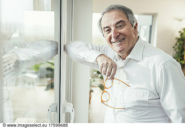 Smiling senior man with eyeglasses standing by window at home