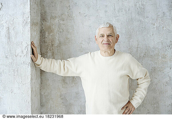 Smiling senior man with hand on hip standing near wall