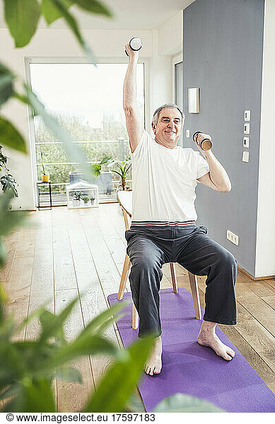Smiling senior man with hand raised holding dumbbell on chair at home