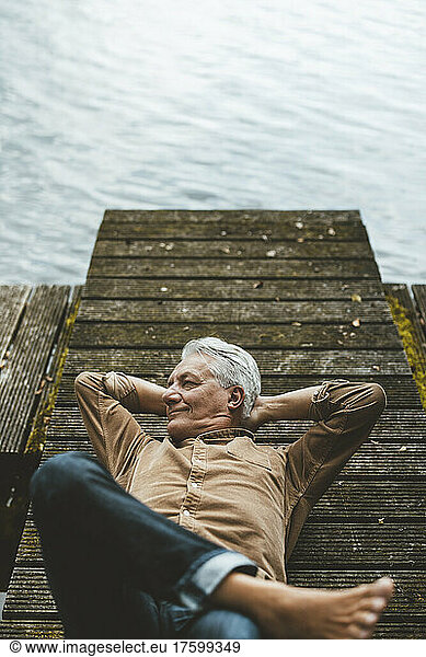 Smiling senior man with hands behind head lying on jetty by lake