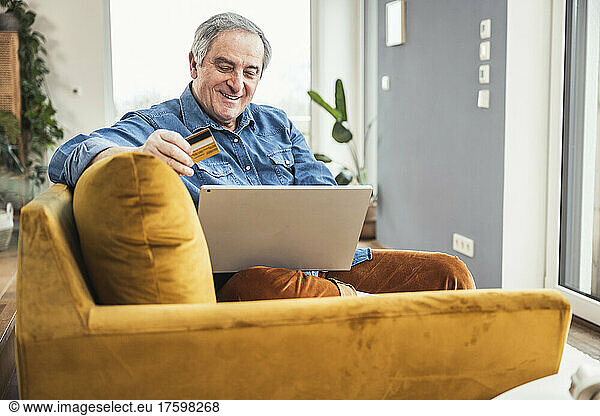 Smiling senior man with laptop and credit card sitting on sofa in living room