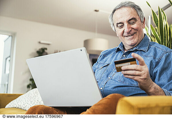 Smiling senior man with laptop looking at credit card in living room at home