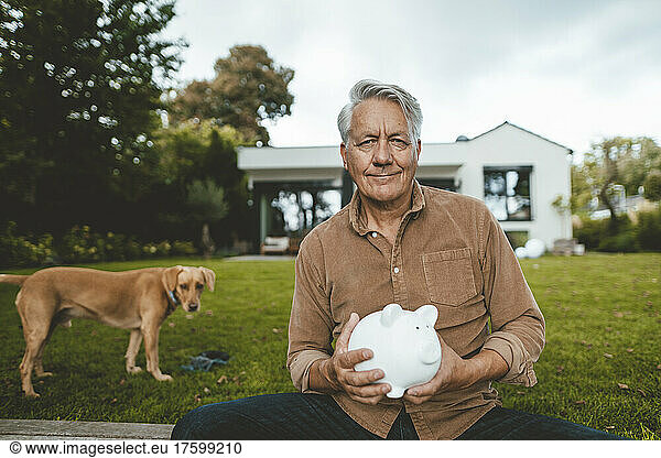 Smiling senior man with piggy bank sitting by dog at backyard