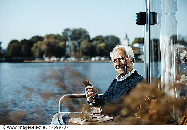 Smiling senior man with smart phone sitting at houseboat on sunny day