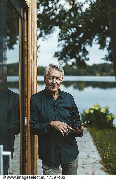 Smiling senior man with tablet PC standing by glass wall at backyard