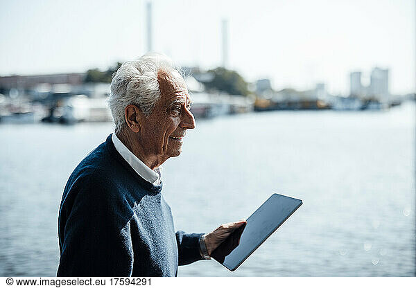 Smiling senior man with white hair holding tablet PC on sunny day by the water