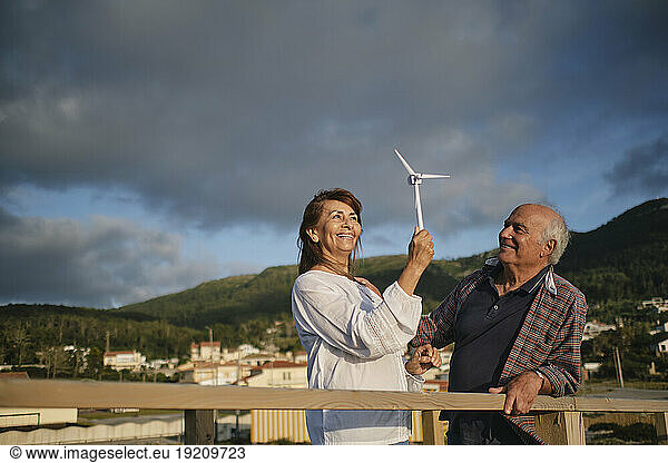 Smiling senior man with woman holding wind turbine under cloudy sky