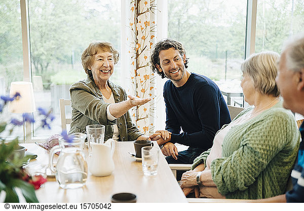 Smiling senior woman gesturing towards friends while sitting with grandson at nursing home