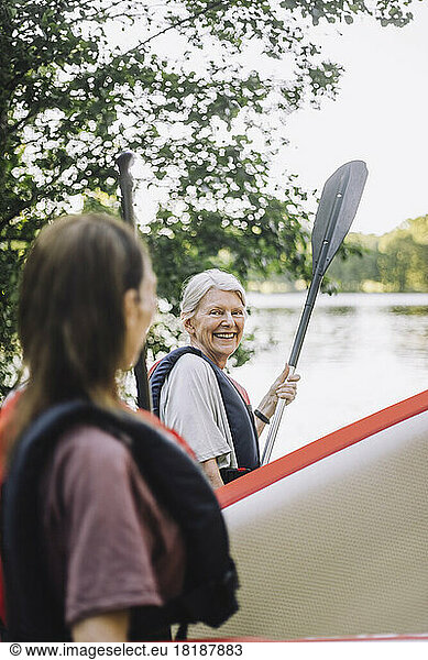 Smiling senior woman holding paddleboard and oar talking with female friend