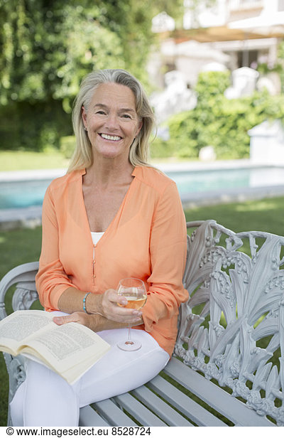 Smiling senior woman reading book and drinking wine in backyard