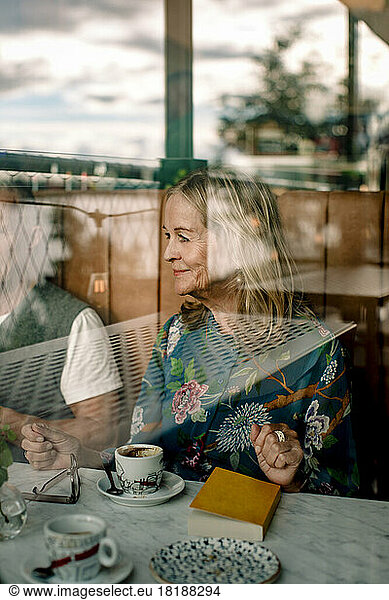 Smiling senior woman sitting with book by male friend in cafe seen through glass