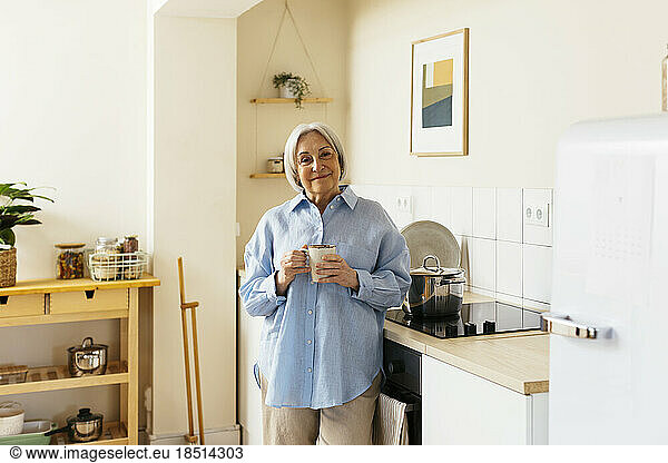 Smiling senior woman standing with cup in kitchen