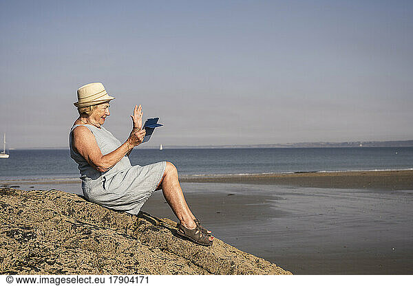 Smiling senior woman waving hand on video call sitting at beach