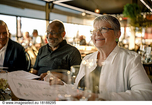 Smiling senior woman wearing eyeglasses sitting with menu card by men in restaurant