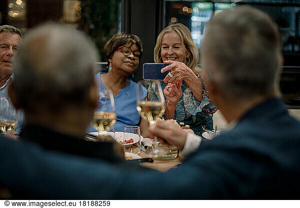 Smiling senior women photographing male friends holding wineglasses in restaurant