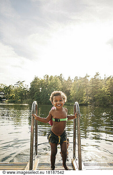 Smiling shirtless boy holding railing while standing on jetty near lake during vacation