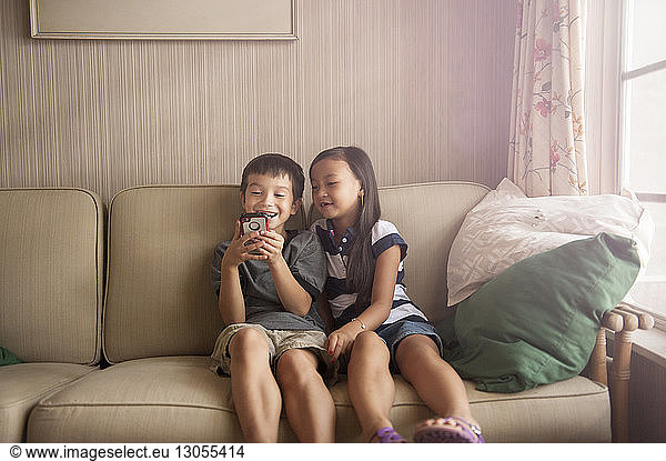 Smiling siblings using phone while relaxing on sofa