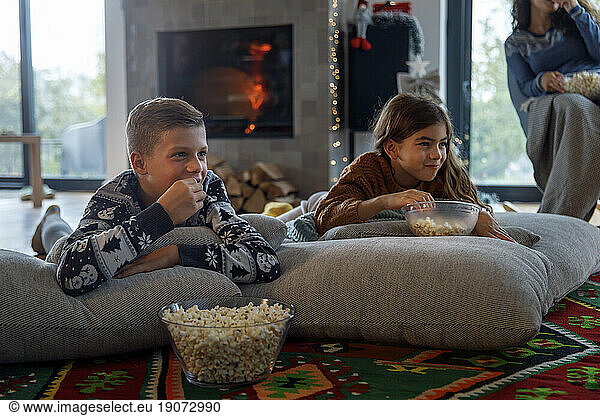 Smiling siblings watching TV with mother and eating popcorn at home
